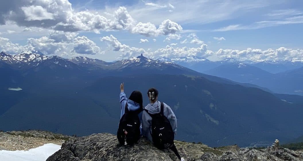 Dois alunos sentados no alto de uma montanha com picos nevados ao fundo
